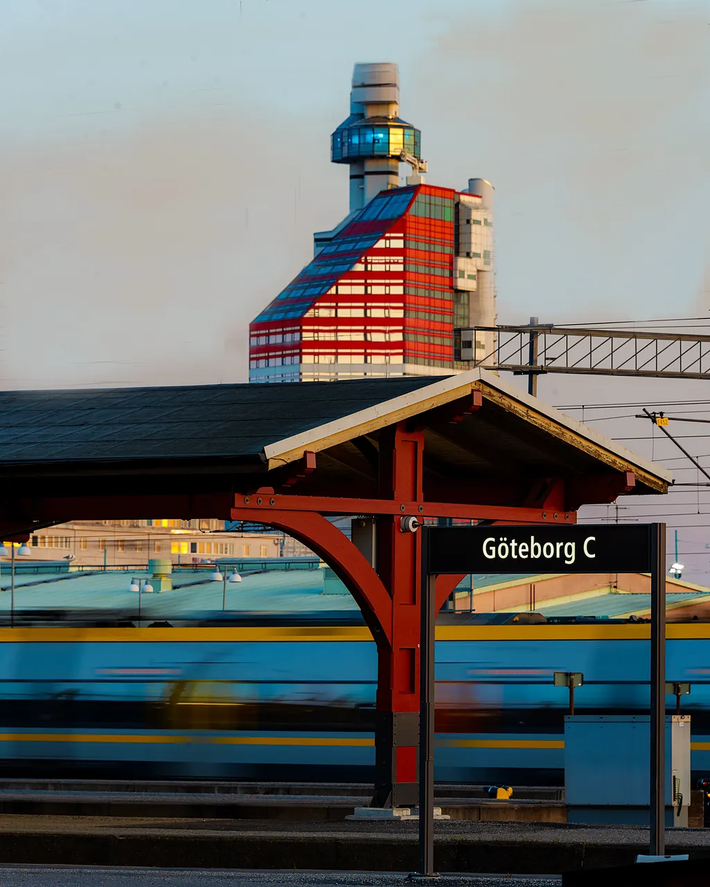 The Göteborg C platform sign at Gothenburg Central Station, with a train passing in a blur and the red-and-white Lilla Bommen tower behind it